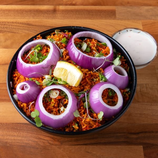 A black bowl of orange-red rice, garnished with red onion rings, green herbs, and a lemon wedge.