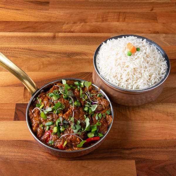 Indian curry in a brass pan with a side of white rice, garnished with peas and carrots.