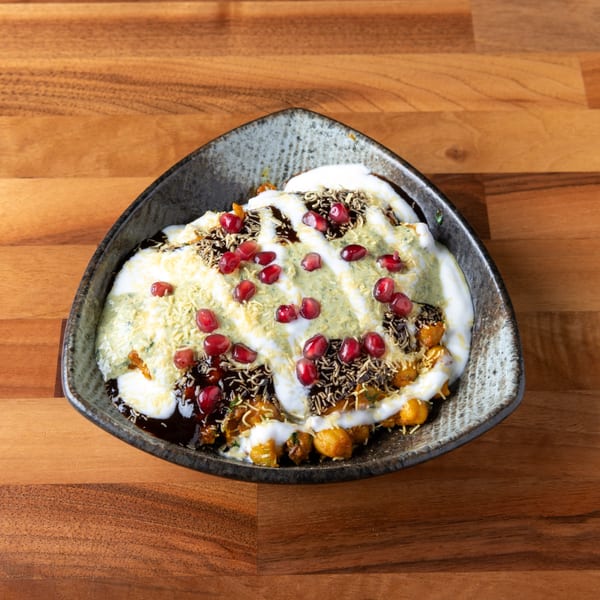 A dark, triangular bowl filled with a colorful dish, topped with white sauce, green chutney, and pomegranate seeds.