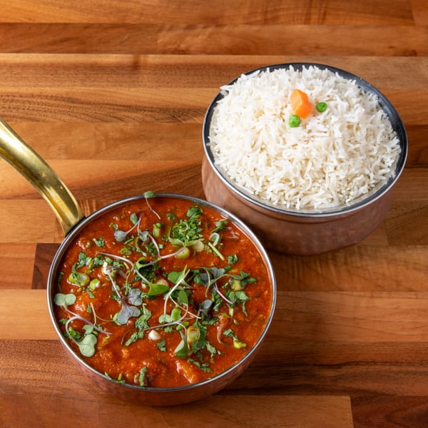 A copper pot of red curry with herbs and a bowl of white rice with peas and carrots.