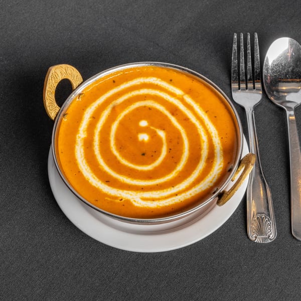 Indian curry in a metal bowl with decorative handles, garnished with a cream swirl, beside silverware.