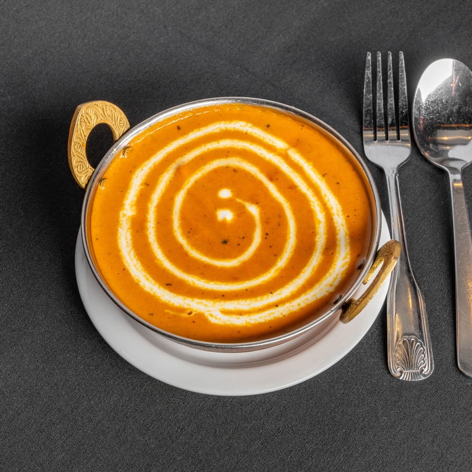 Indian curry in a metal bowl with decorative handles, garnished with a cream swirl, beside silverware.