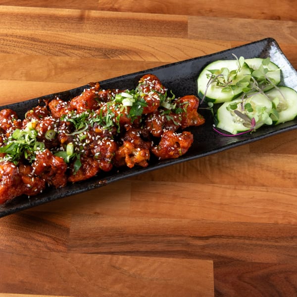 Spicy Korean fried chicken bites with sesame seeds, green onions, and cucumber slices on a black plate.