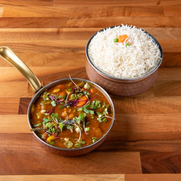 A copper pot of vegetable curry with microgreens and a bowl of white rice on a wooden table.