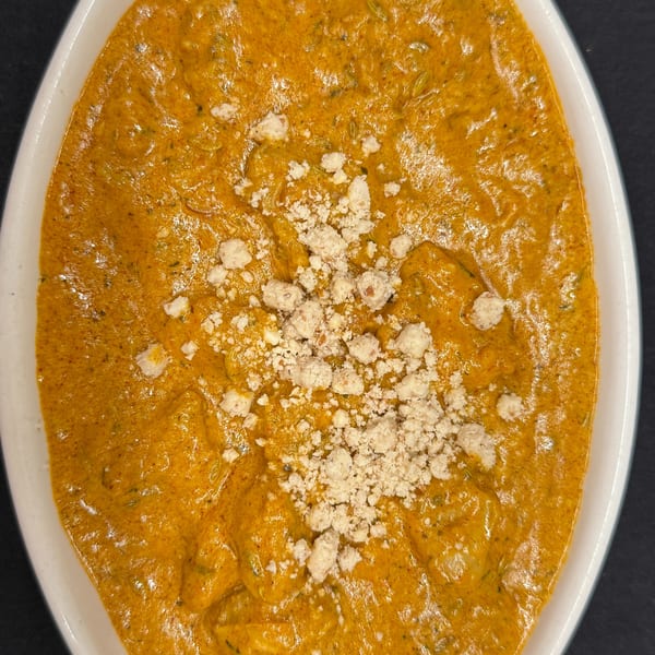 Close-up of a rich orange Indian curry dish, topped with crumbled white cheese, in a white oval bowl.