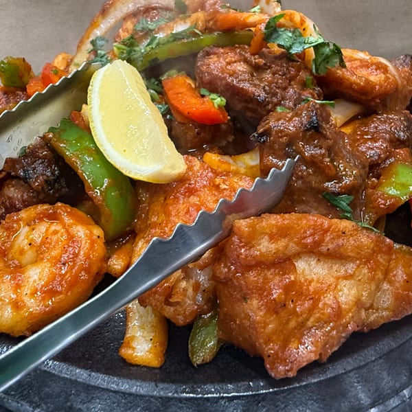 Sizzling Indian mixed grill with chicken, shrimp, vegetables, and a lemon wedge, served with tongs.