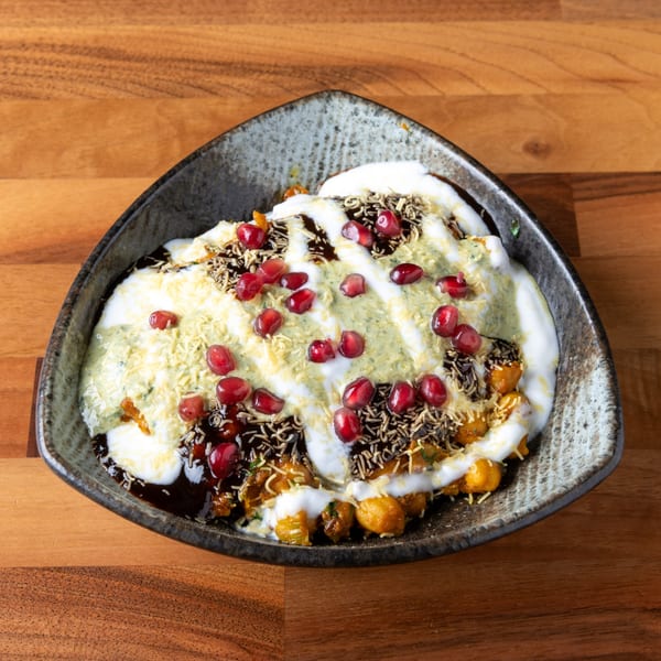 A dark, triangular bowl filled with Indian chaat, topped with yogurt, chutneys, sev, and pomegranate seeds.