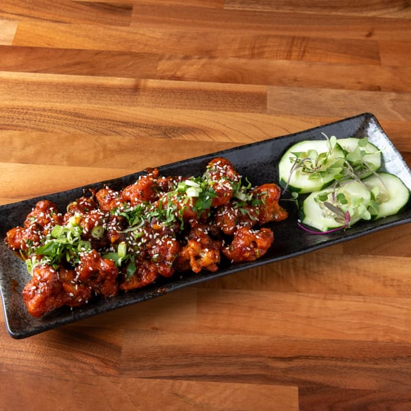 Spicy Korean fried chicken bites with sesame seeds, green onions, and cucumber slices on a black plate.