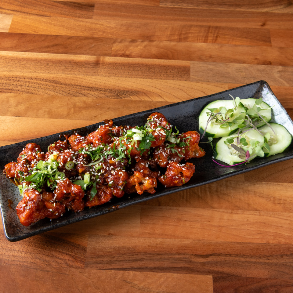 Spicy Korean fried chicken bites with sesame seeds, green onions, and cucumber slices on a black plate.