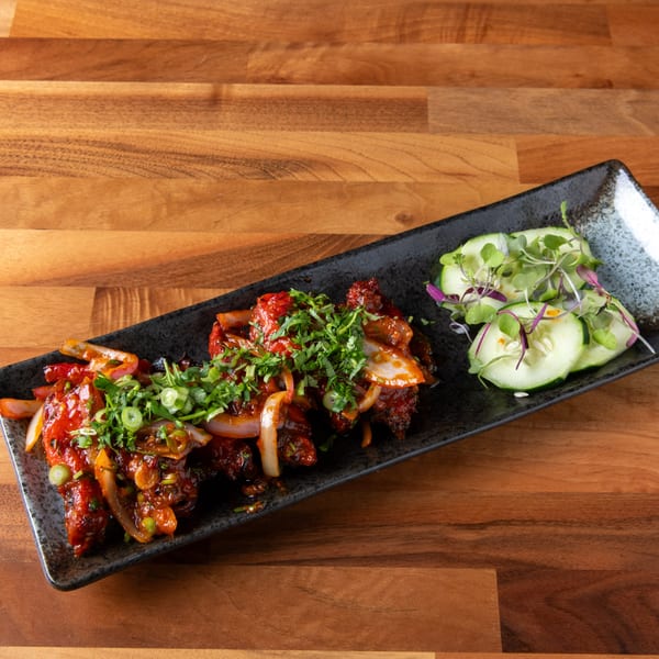 Spicy red chicken dish with onions and cilantro, served with cucumber slices on a black plate.